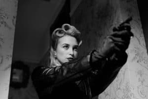 Black and white image of a woman in vintage attire, pointing a stun gun. She wears a leather jacket and has styled hair, standing against a patterned wallpaper background.