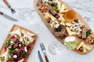 A charcuterie board with cheese, nuts, honey, and pate on a marble table next to a wooden board with meat, arugula, and cheese. Plates, glasses, and cutlery are also visible.