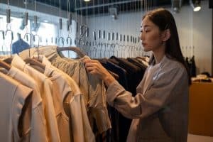 A woman is browsing through clothes on hangers in a clothing store. She is touching a white blouse while looking at the garments.