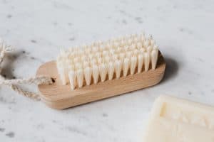 A wooden hand brush with white bristles and rope is placed on a marble surface next to a bar of soap.