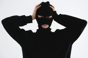 Person wearing a black balaclava and black clothing, holding their head with both hands against a plain background.