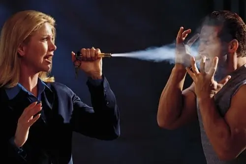 A woman sprays a substance into the face of a man using a small spray device. The man appears to be in distress, holding his hands near his face.