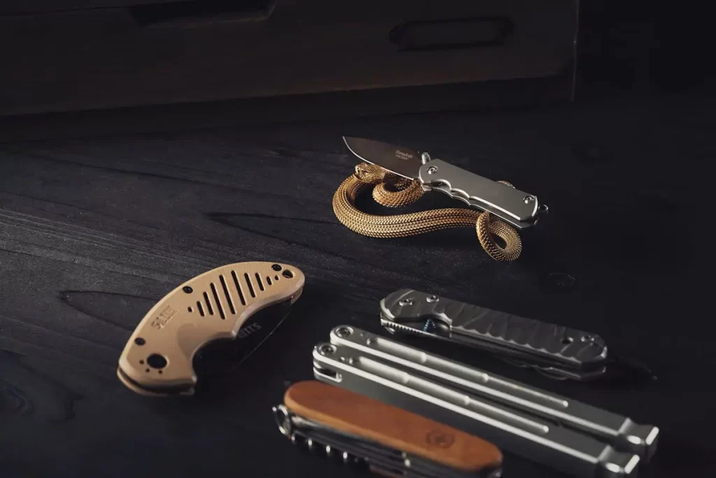 A collection of sleek, modern pocket knives and a multi-tool are arranged on a dark surface, with a small bronze-colored snake encircling one of the knives.