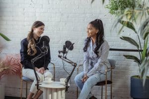 Two women seated and speaking into microphones in a bright room with plants, possibly recording a podcast. Two cups are on the small table between them.