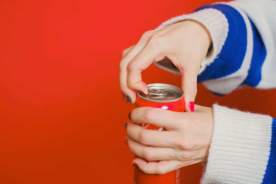 A person wearing a white and blue sweater is opening a red soda can against a red background.