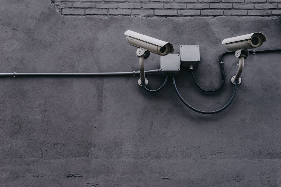 Two security cameras are mounted on a gray wall, connected by wires and mounted on brackets.