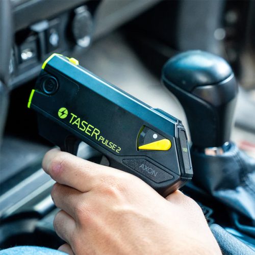 In a vehicle near the gear shift, a hand grips the sleek black and yellow Taser Pulse 2 self-defense device.